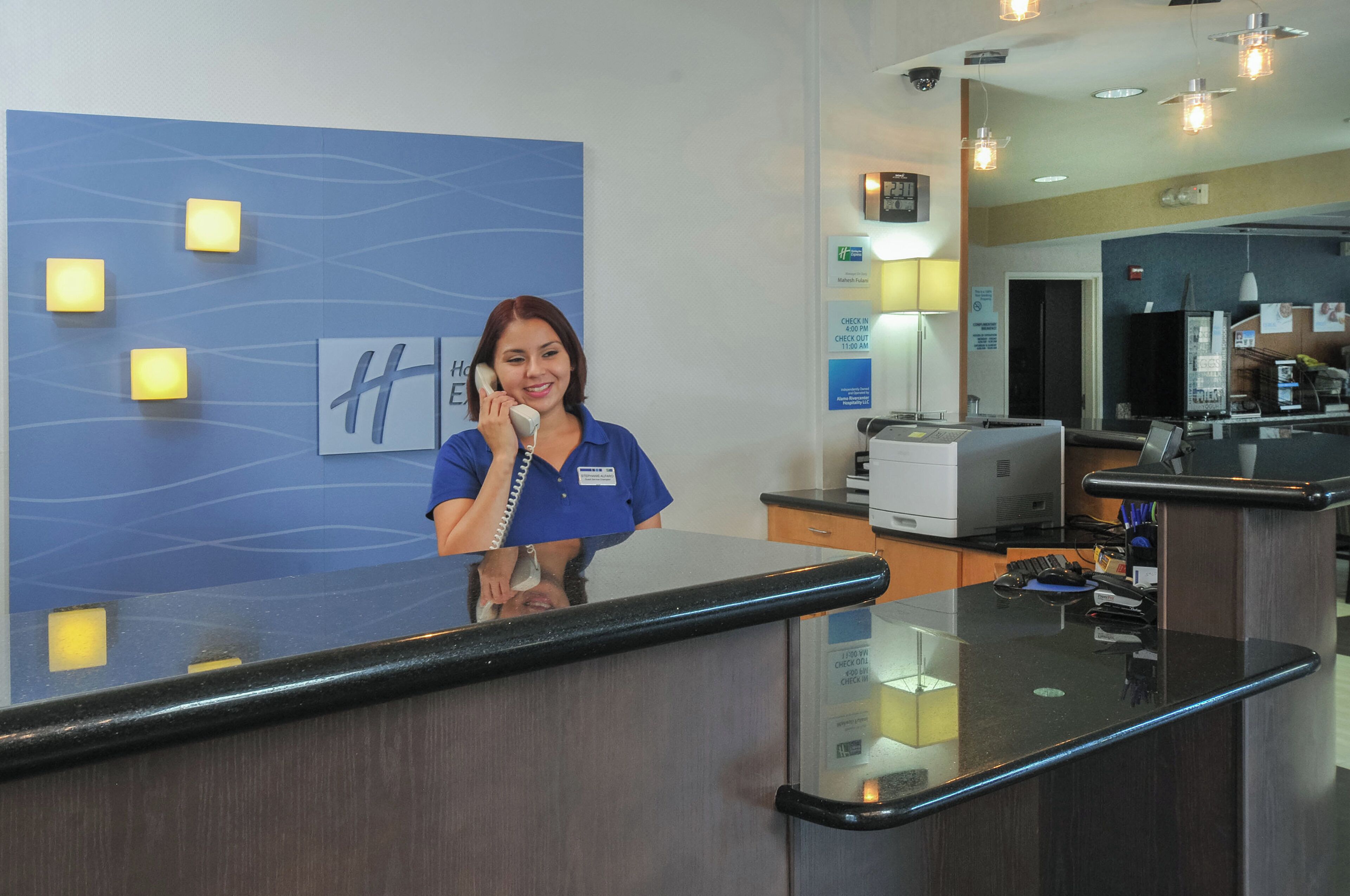 Zona de recepción de un hotel con recepcionista, un mostrador de recepción, un teléfono en la pared y un reloj digital.