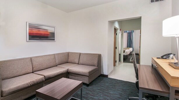 Une chambre d’hôtel moderne avec un canapé d’angle, une petite table, un bureau avec une lampe et un tableau encadré au mur.