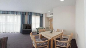 Et hotelværelse med et spisebord, fire stole, et fjernsyn og et stort vindue med gardiner.