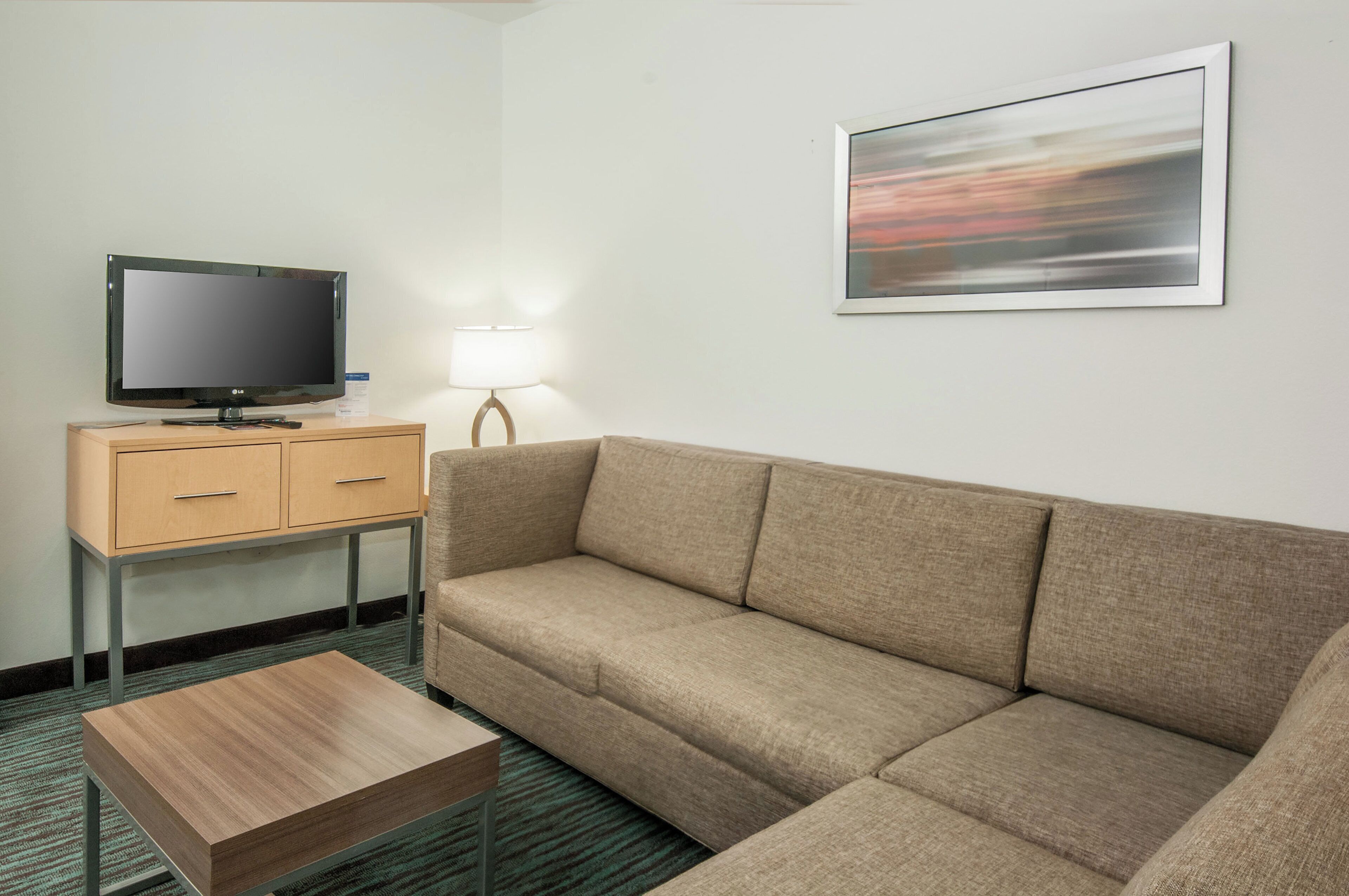 A hotel room with a sofa, a television on a wooden cabinet, a lamp, and a framed picture on the wall.