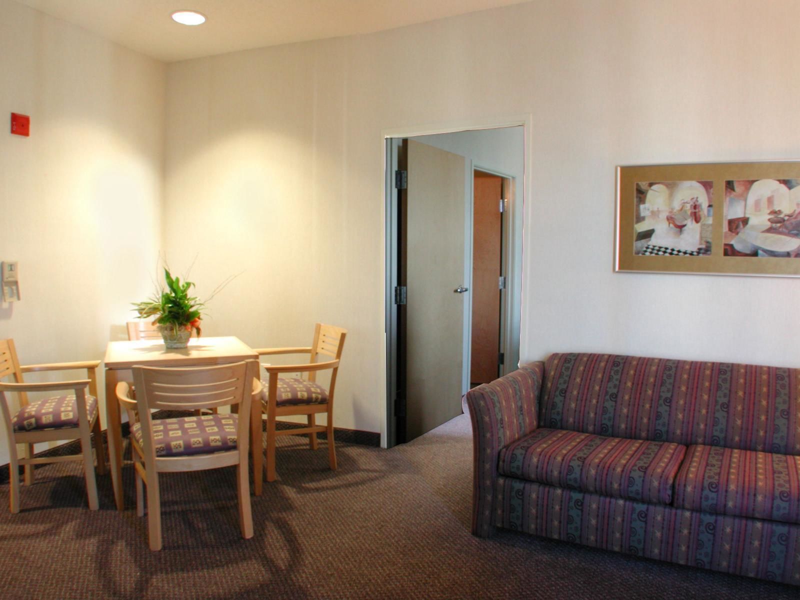 Habitación de hotel con zona de comedor que incluye una mesa y cuatro sillas, un sofá y un cuadro en la pared.