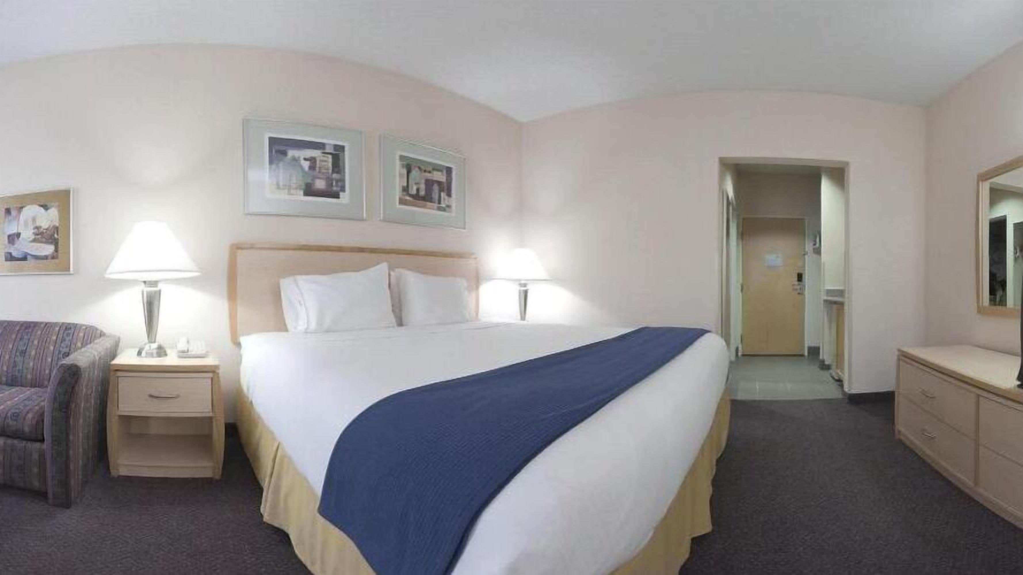 Habitación de hotel con cama, dos mesitas de noche con lámparas, una silla, cómoda y una puerta que da a otra habitación.