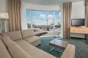 Ein Hotelzimmer mit großem Fenster, einer Couch, einem kleinen Tisch, einem Fernseher und einem Schreibtisch mit Schubladen.