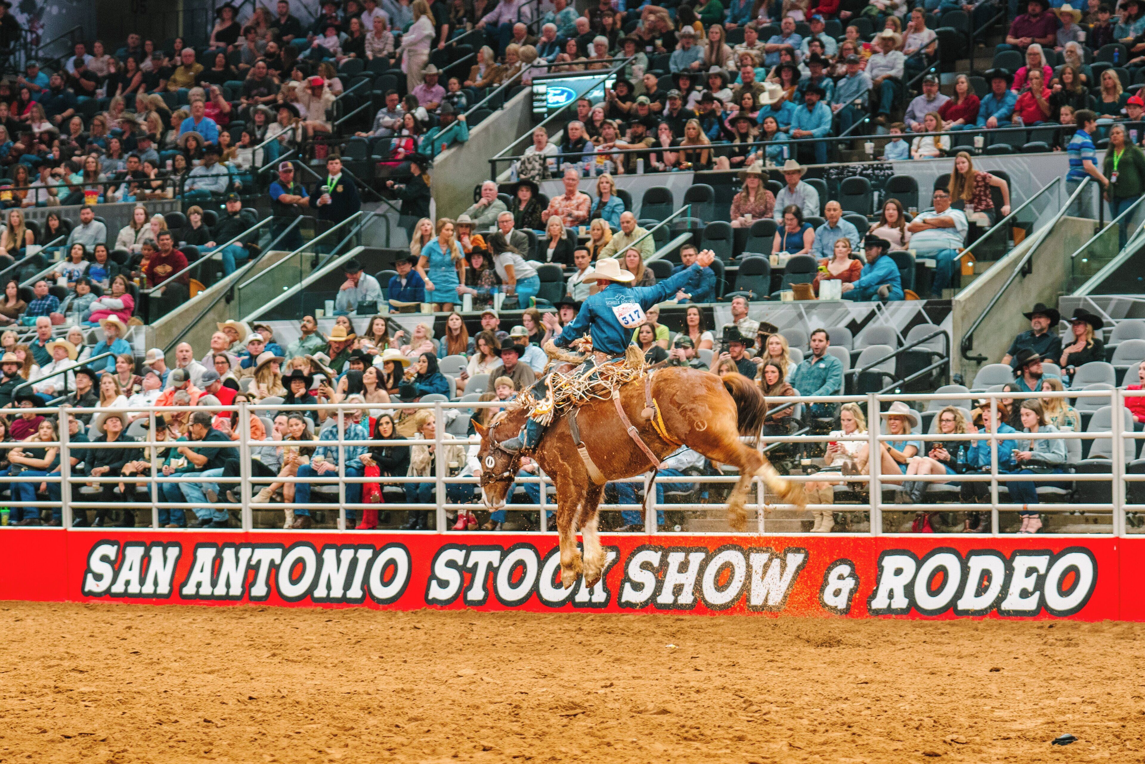 A rodeo event with a rider on a bucking horse, spectators in the stands, and a sign reading "SAN ANTONIO STOCK SHOW & RODEO".