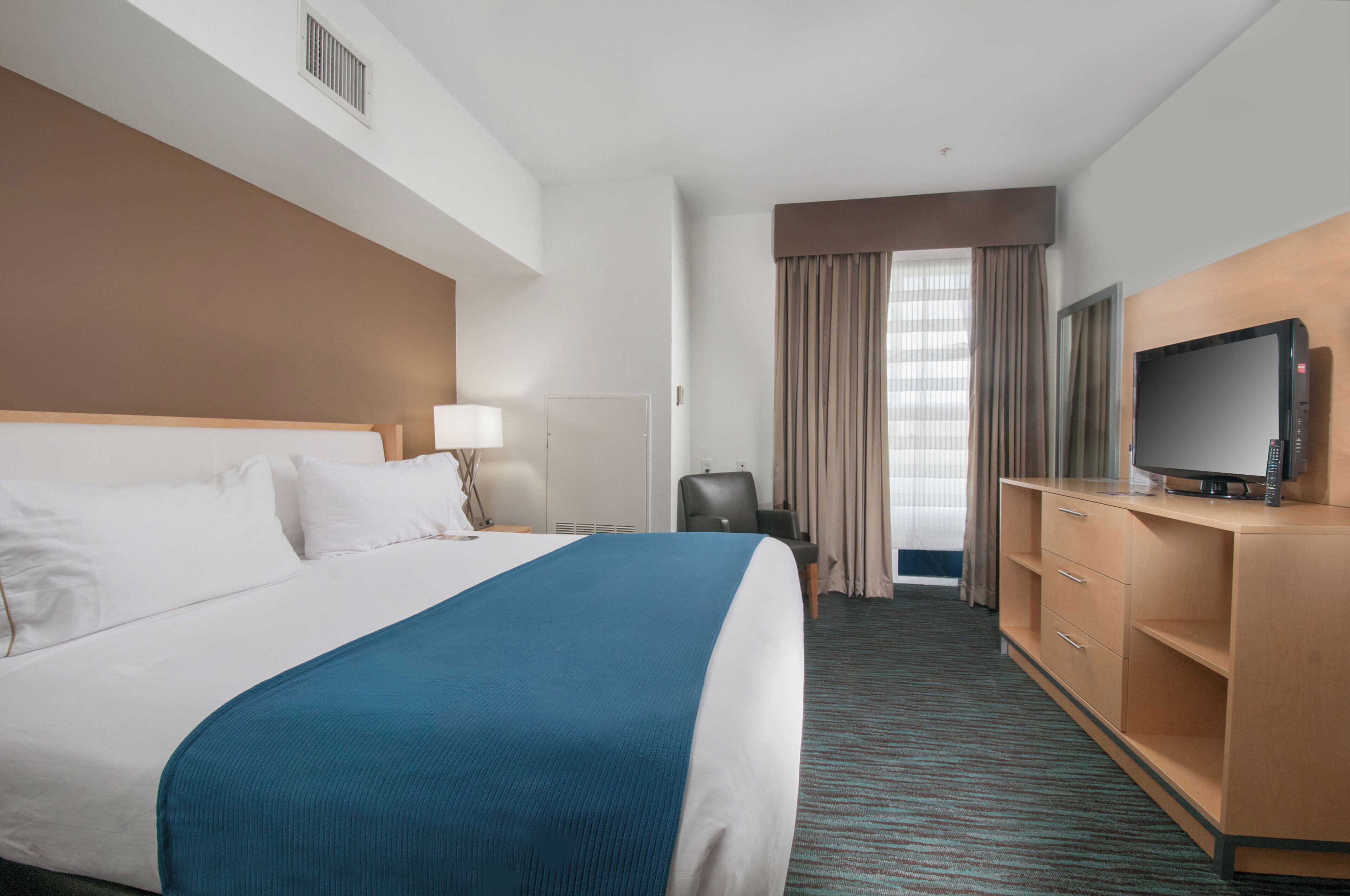 Habitación de hotel con una cama grande, un televisor sobre un mueble de madera, un escritorio con lámpara y vistas al exterior a través de una ventana.