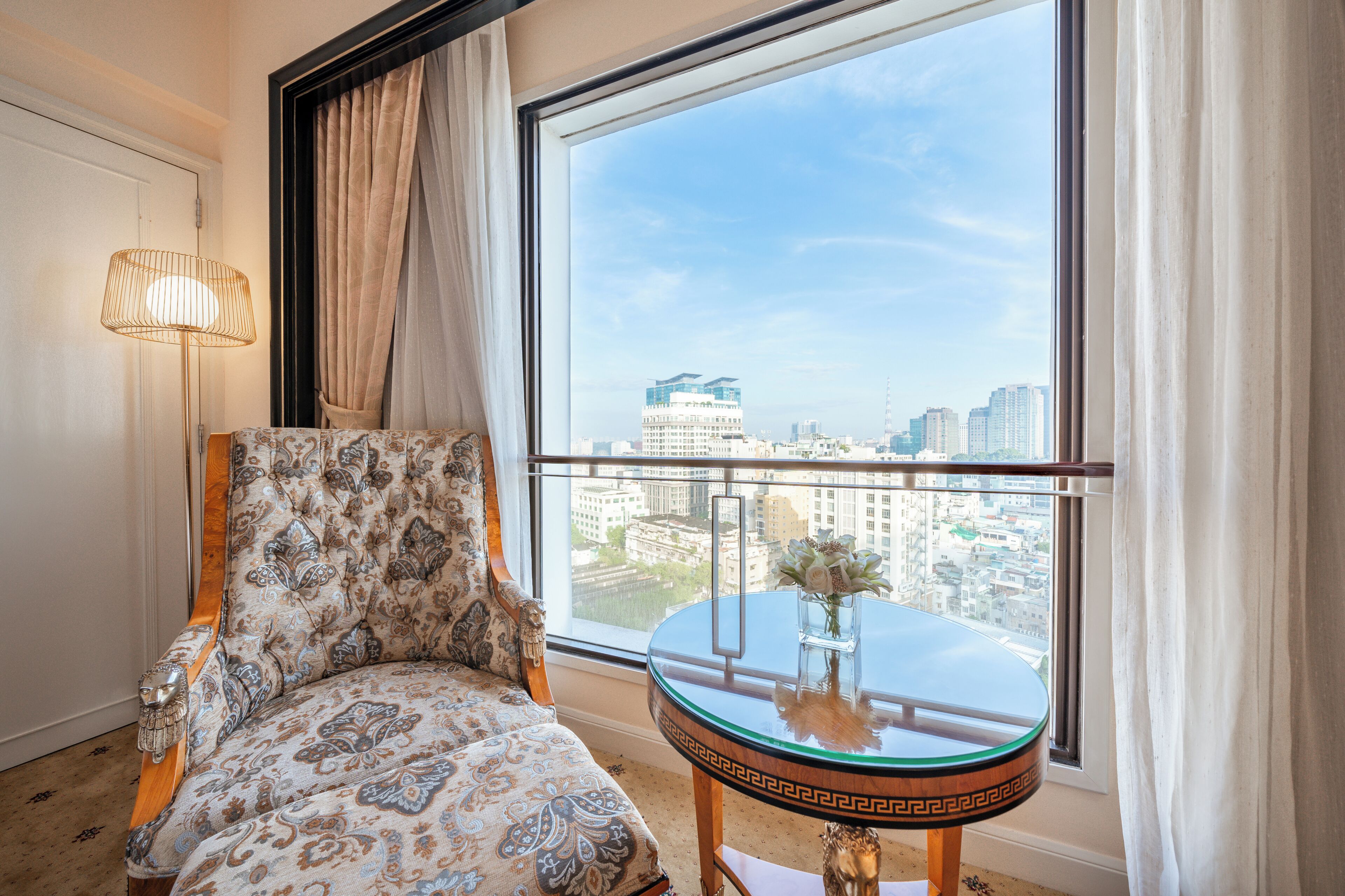 A room with a large window, a patterned sofa, a small round table with a vase, and a floor lamp.