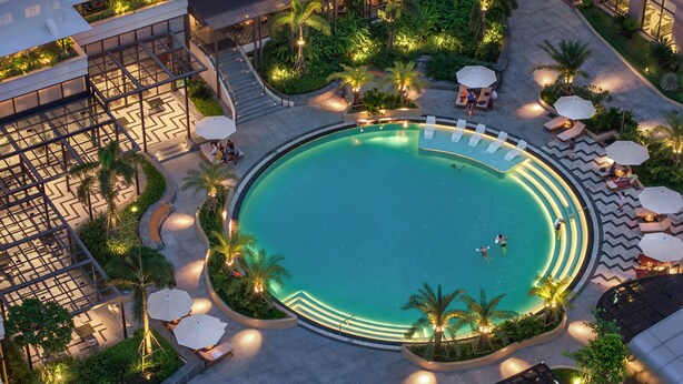 An aerial view of a resort with a central swimming pool surrounded by lounge areas, palm trees, and a covered walkway. - Lotte Hotel Saigon (Ho Chi Minh City)