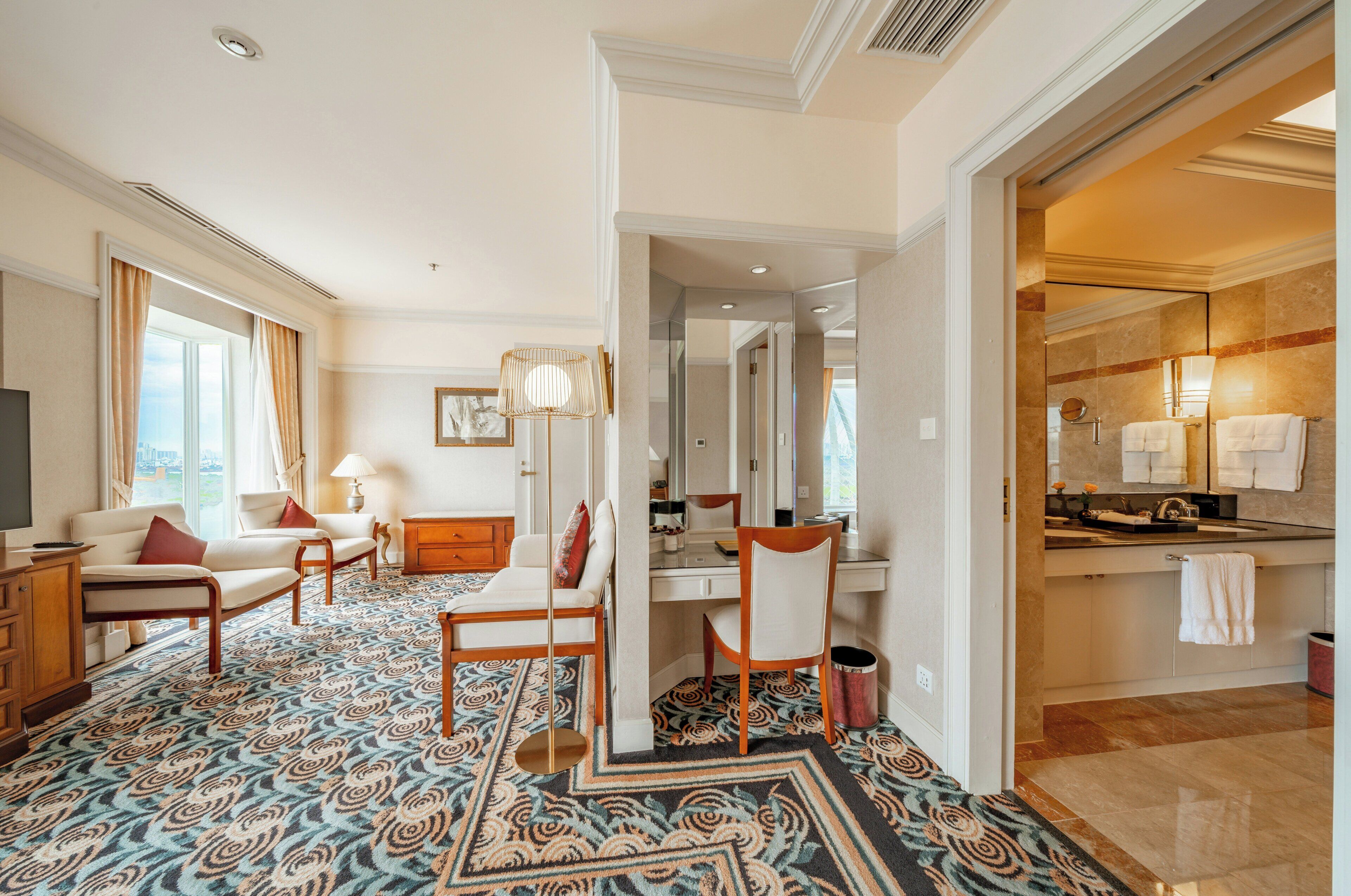 Habitación de hotel con alfombra estampada, un sofá, un escritorio, una silla, una lámpara, un espejo y un baño con lavamanos y toallero.