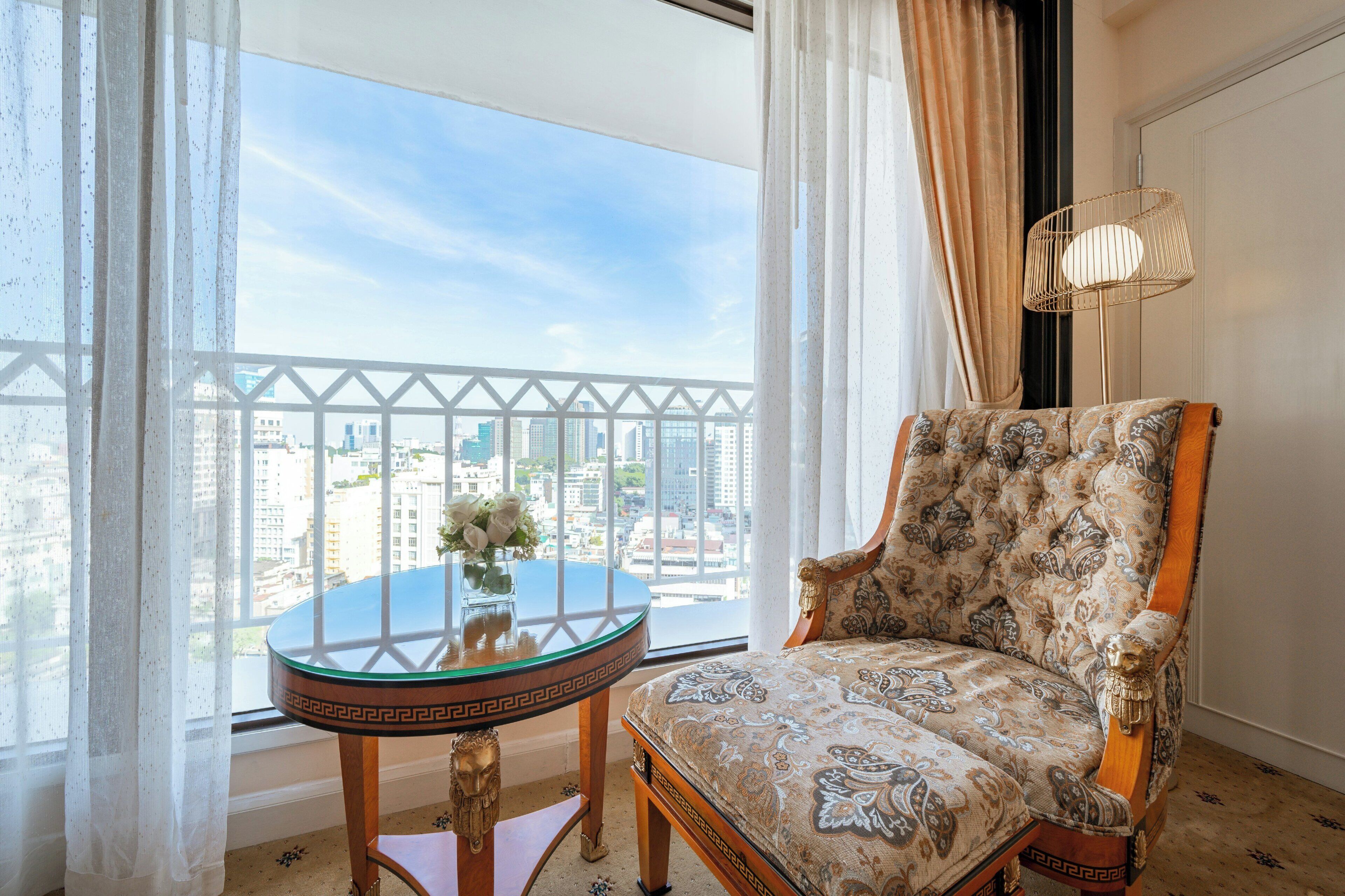 A balcony with a view of a cityscape, a round glass table with a vase of flowers, and a patterned armchair.
