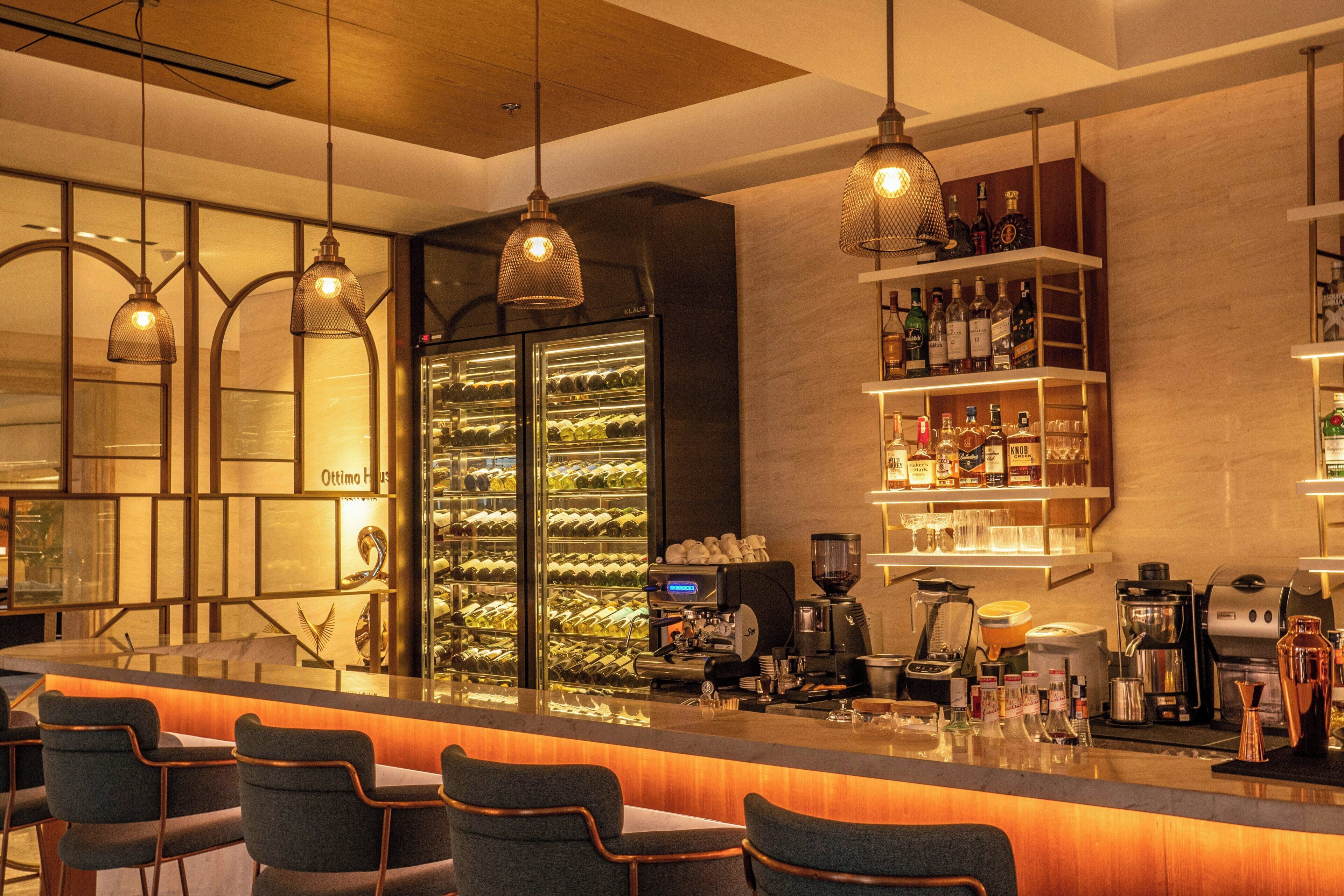 Una zona de bar con mostrador, taburetes, un enfriador de vino y una variedad de botellas y copas.