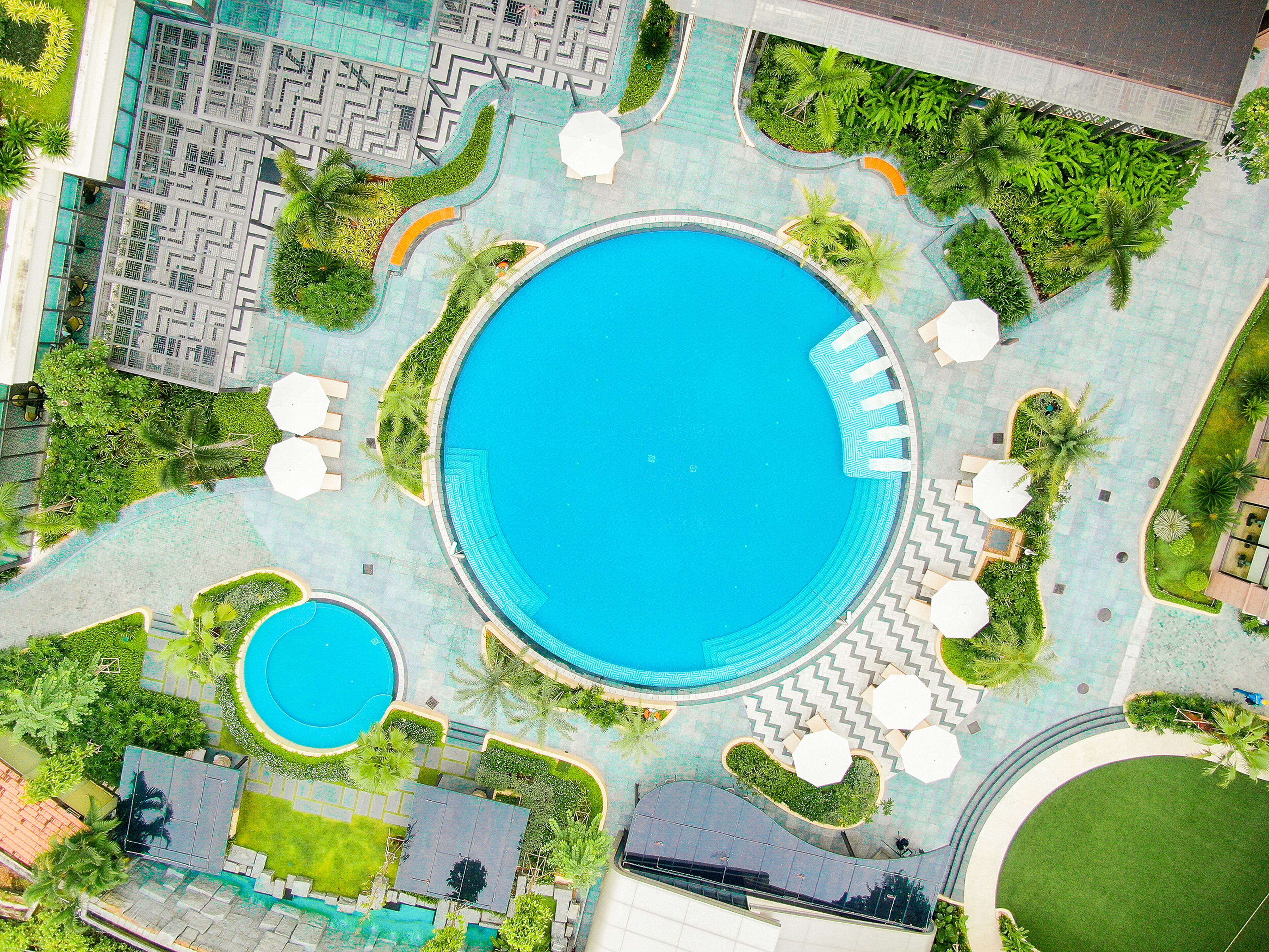 Vista aérea de um resort com uma piscina central grande, uma piscina circular menor e várias áreas de lounge com guarda-sóis.