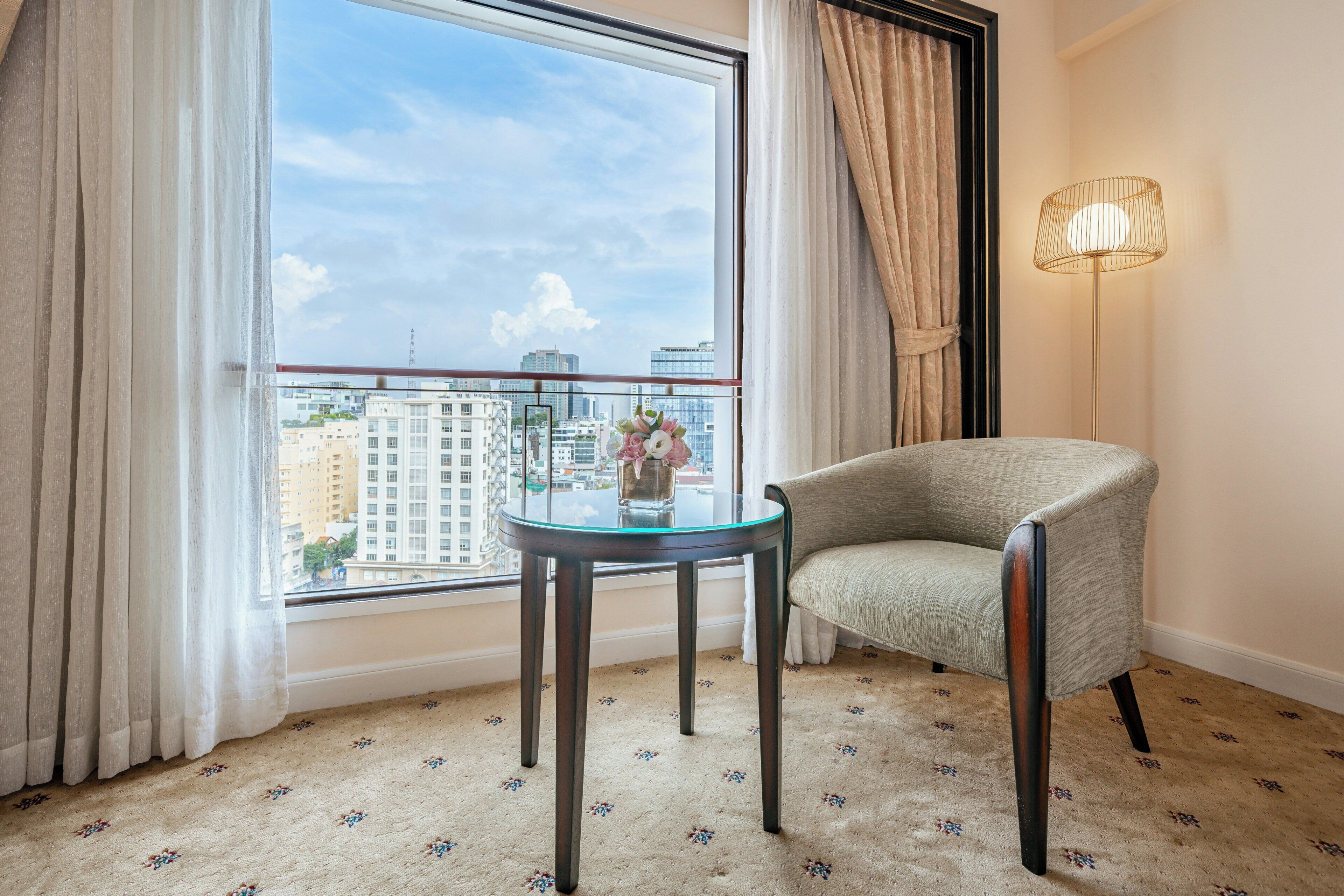 A room with a view of a cityscape, a round glass table, a chair, and a floor lamp.