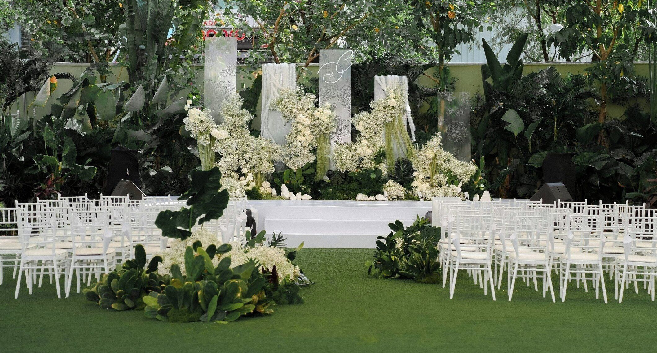 An outdoor wedding venue with white chairs, a stage, and tropical plants.