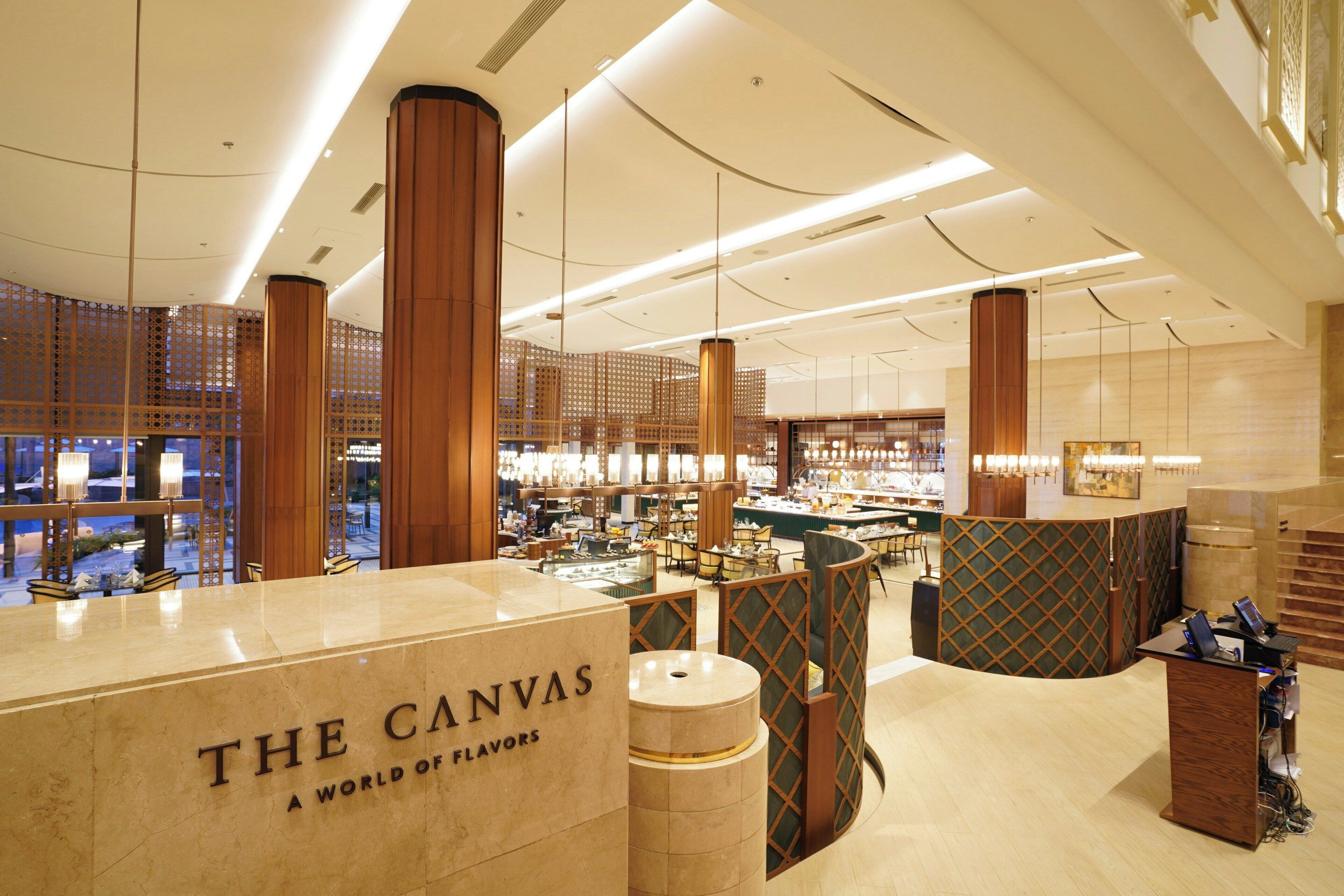 A modern interior with a reception desk labeled 'The Canvas A World of Flavors,' featuring a high ceiling, wooden accents, and a view of the cityscape.
