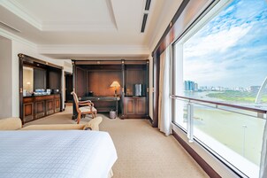 A hotel room with a large bed, a desk with a lamp, a chair, and a view of a cityscape through large windows.