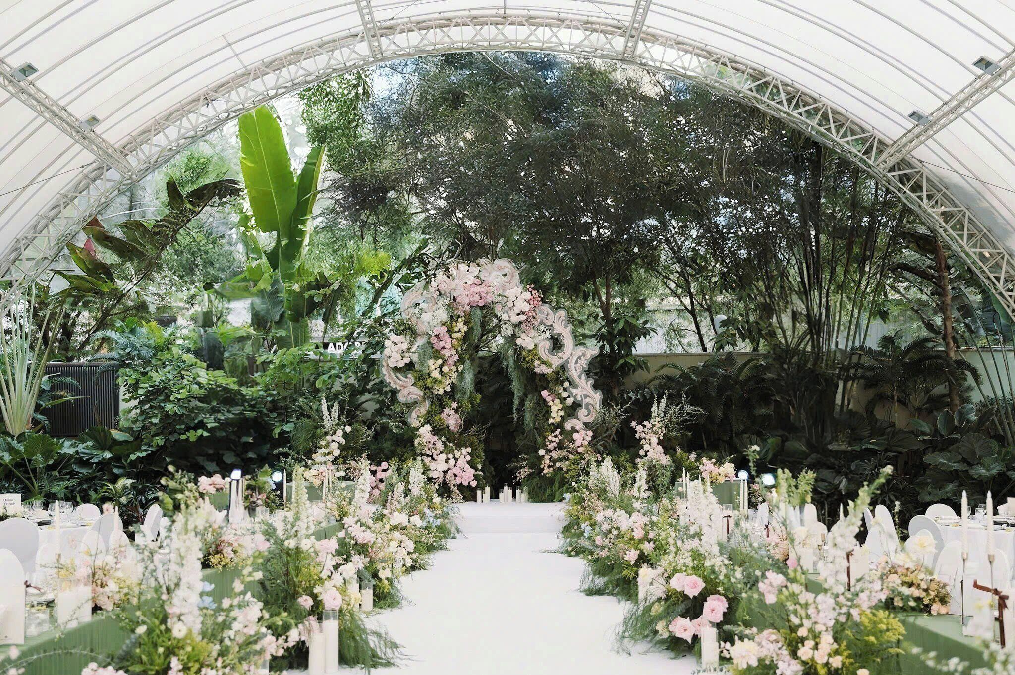 Espaço interno com um corredor branco que leva a um arco floral, rodeado por vegetação exuberante e uma parede de pedra.