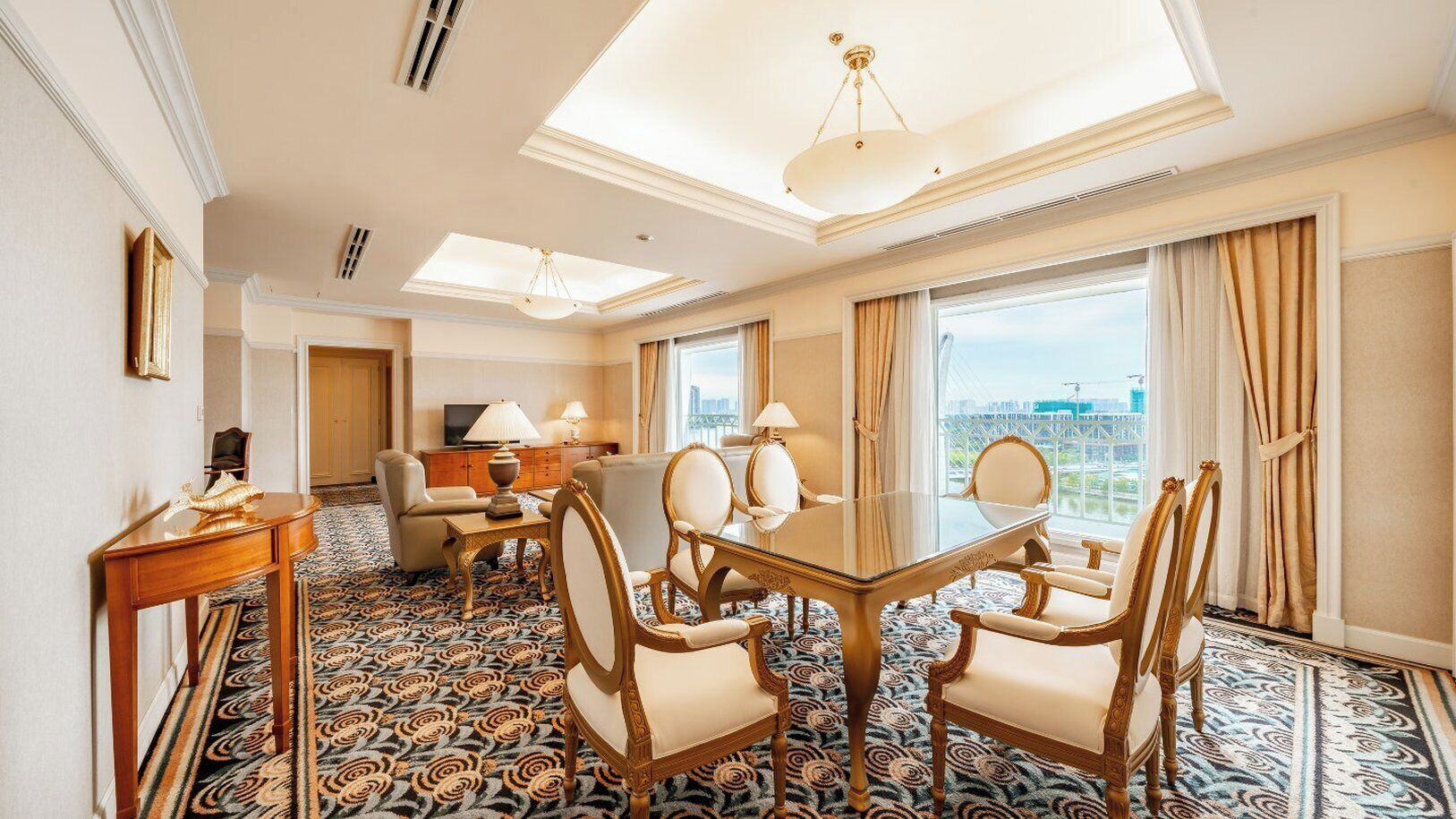A hotel room with a large dining table, four chairs, a sofa, a TV, and a city view.