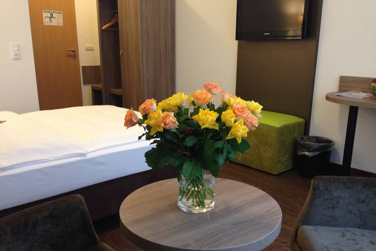 Ein Hotelzimmer mit einem Bett, einem runden Tisch mit Blumenvase, einem grünen Pouf und einem an der Wand befestigten Fernseher.