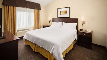 A hotel room with a large bed, two bedside tables, a desk, a chair, a window with curtains, and a wall-mounted picture.