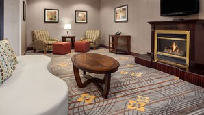 A hotel room with a sofa, two patterned armchairs, a wooden coffee table, a fireplace, and a flat-screen TV mounted on the wall. - Hampton Inn & Suites Detroit/Sterling Heights (Sterling Heights)