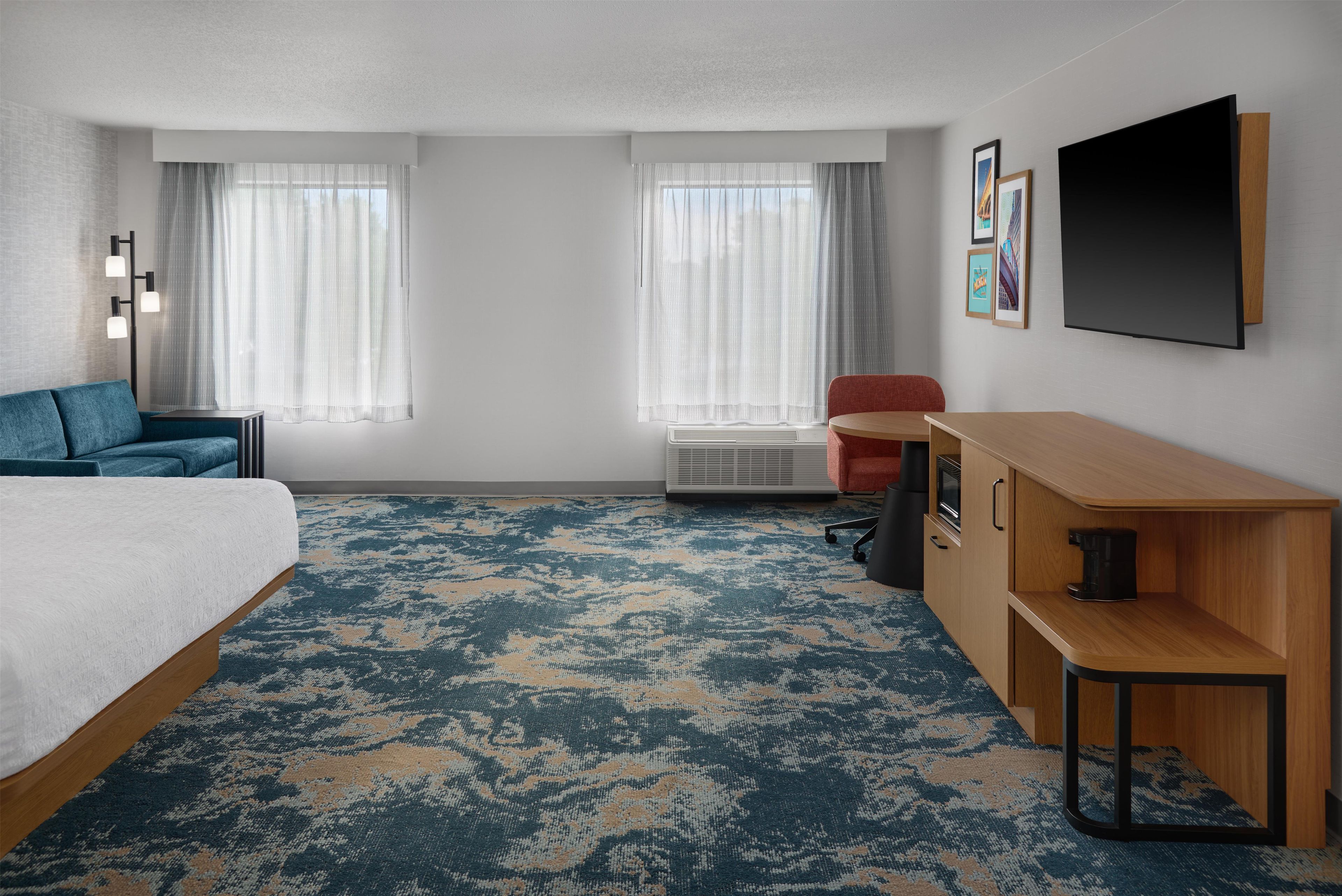 Habitación de hotel con cama, escritorio, silla, sofá y dos ventanas con cortinas.