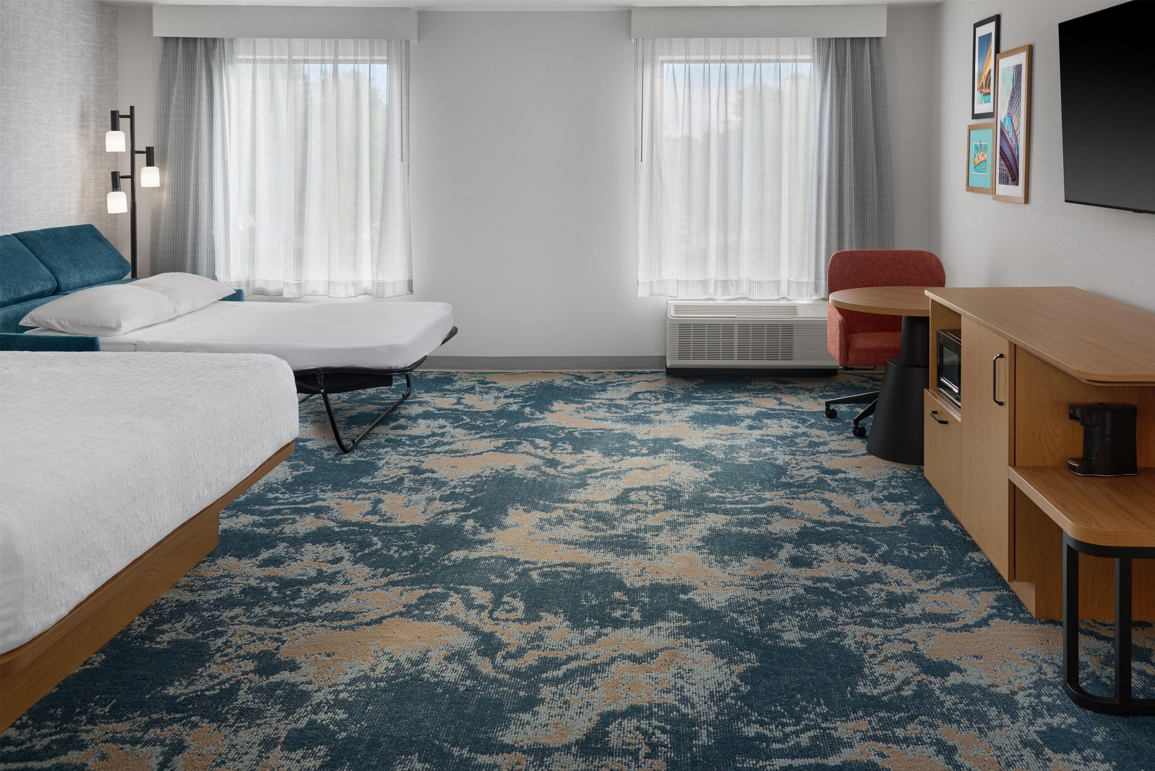 A hotel room with two beds, a desk, a chair, a coffee maker, and a large window with curtains.