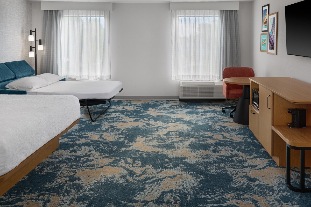 A hotel room with two beds, a desk, a chair, a coffee maker, and a large window with curtains.
