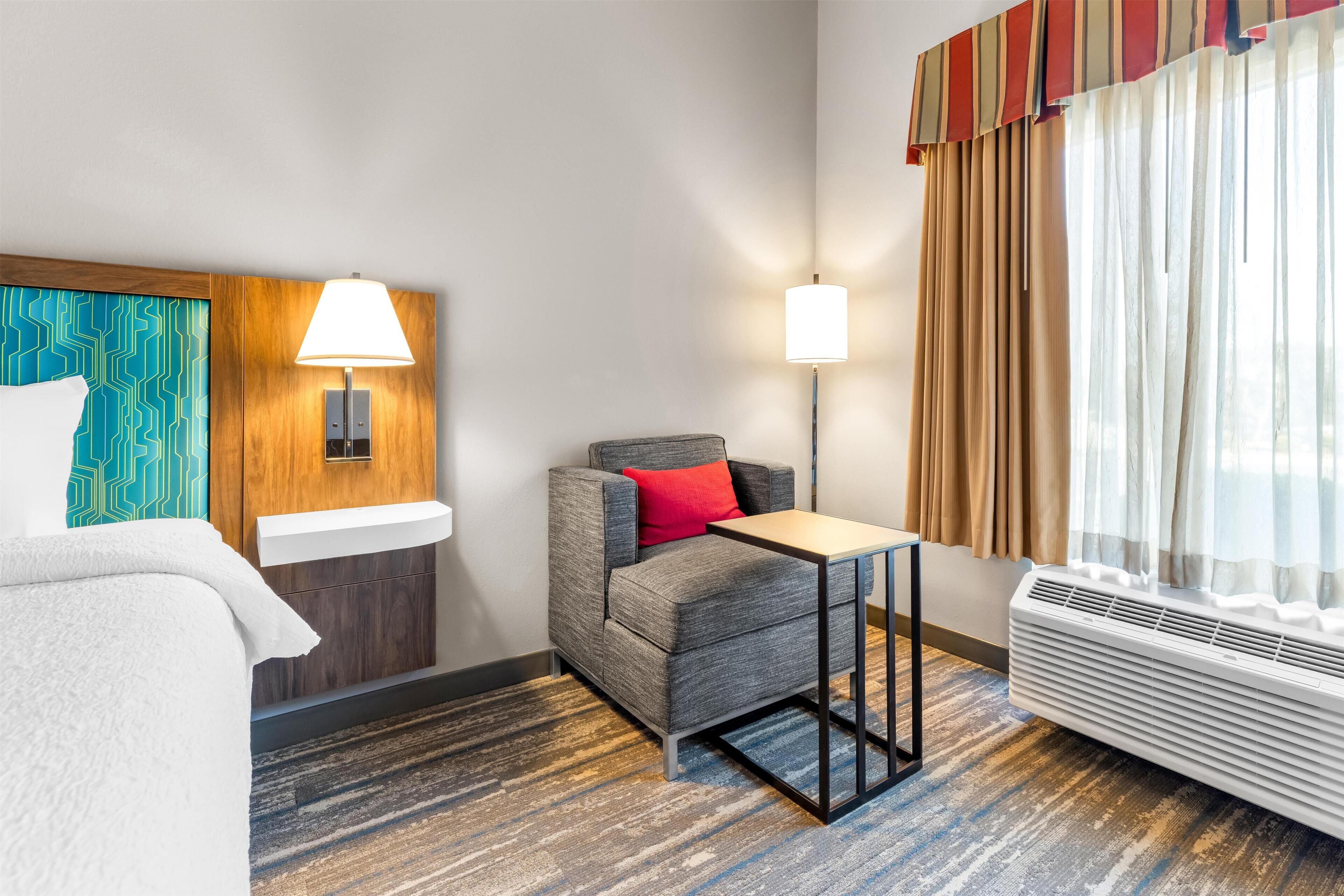 Una habitación de hotel con cama, escritorio, silla, lámpara, calefactor y ventana con cortinas.