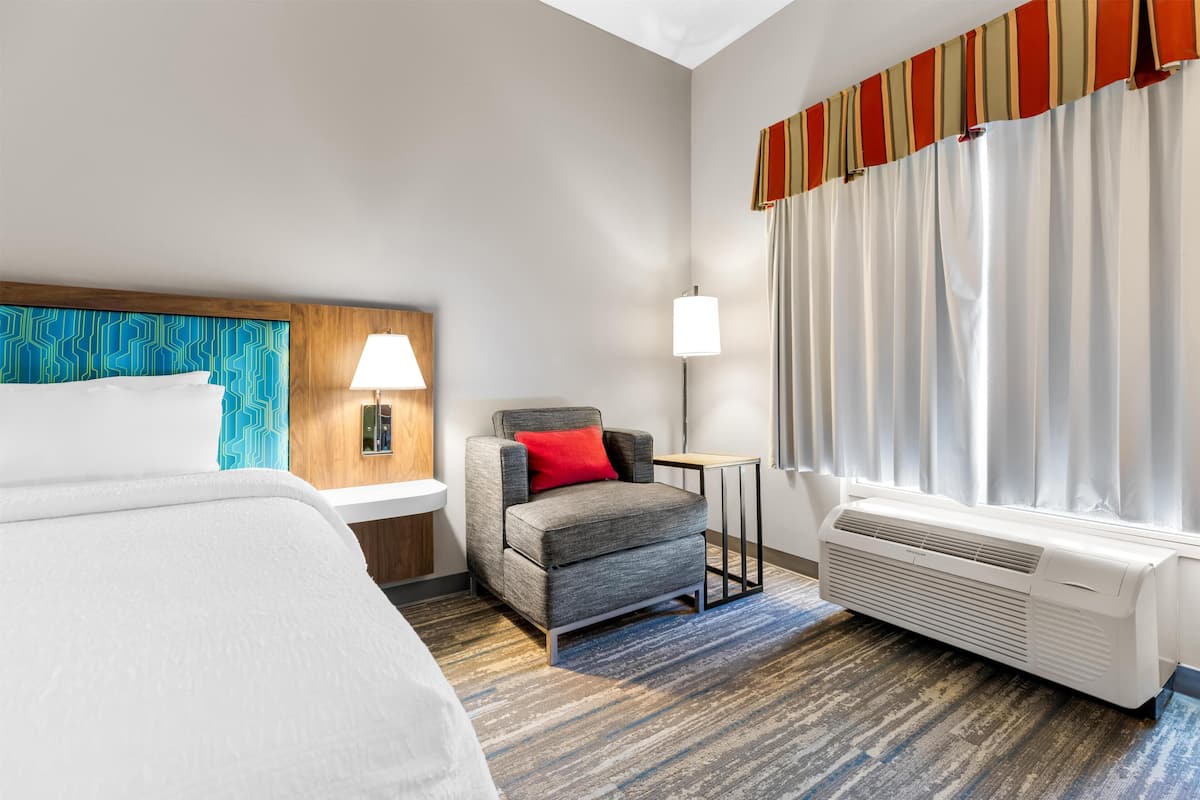 A hotel room with a bed, a chair, a lamp, a heater, and a window with curtains.