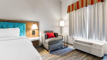 A hotel room with a bed, a chair, a nightstand, a lamp, a window with curtains, and a wall-mounted air conditioner.