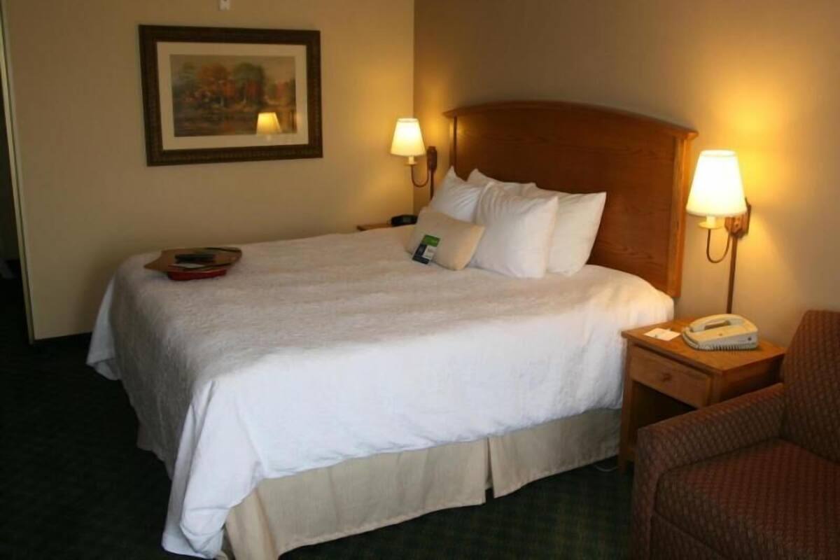 A hotel room with a bed, two bedside lamps, a nightstand with a phone, a framed picture on the wall, and a chair.