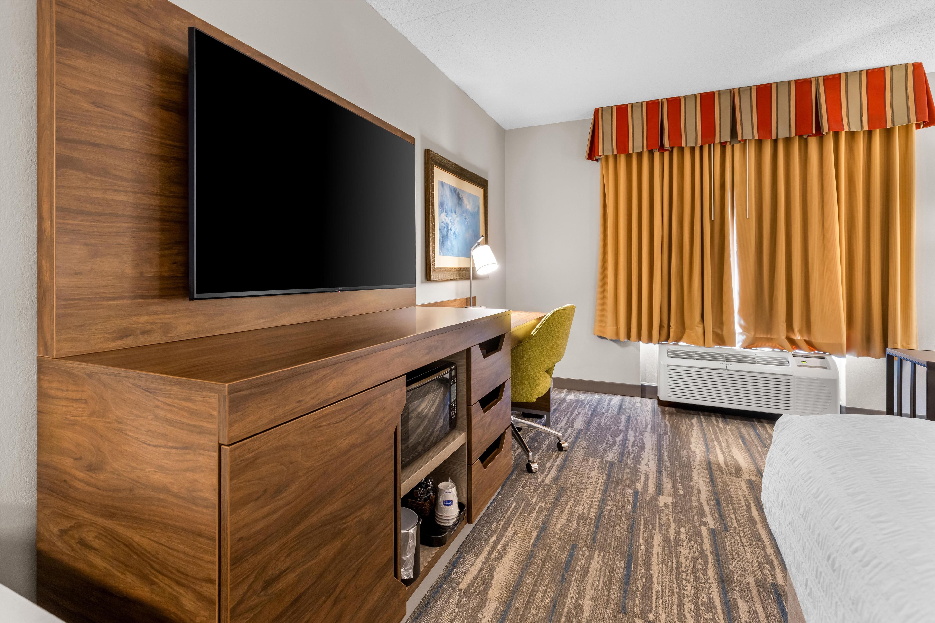 A hotel room with a flat-screen TV mounted on a wooden cabinet, a desk with a chair, a framed picture on the wall, and a window with curtains.