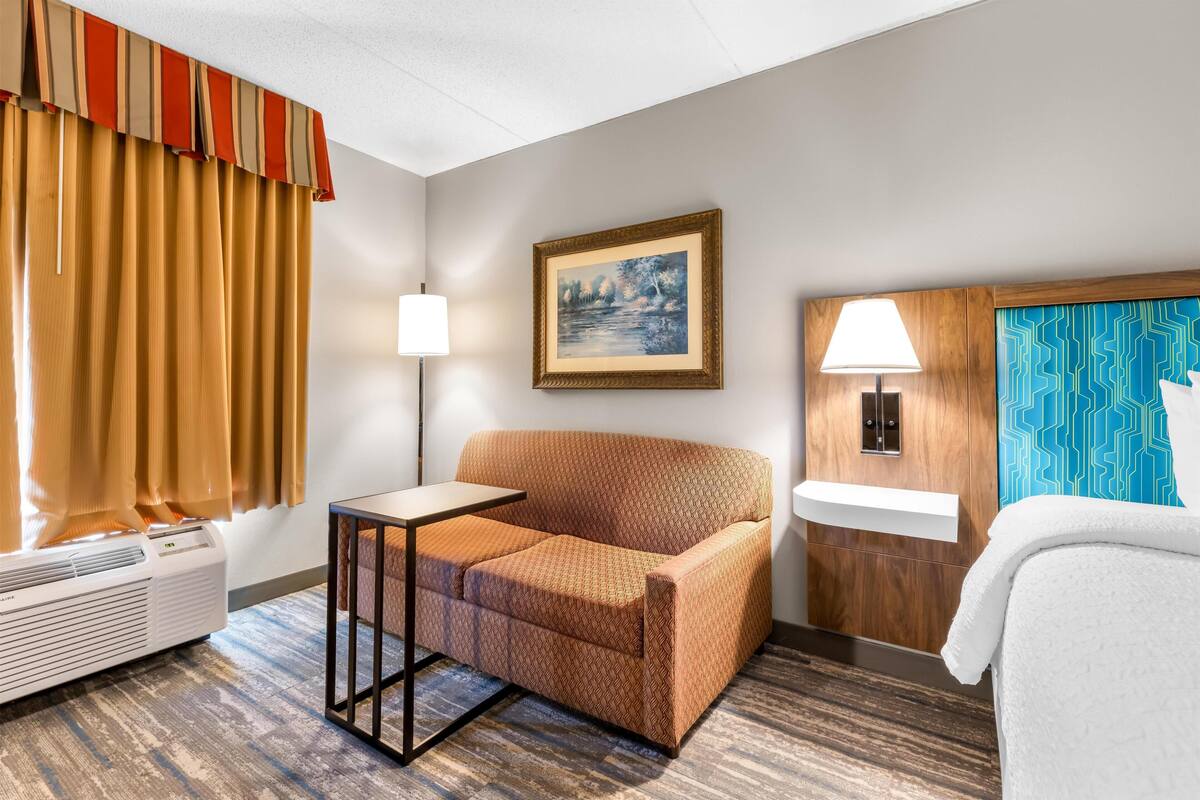 A hotel room with a sofa, a small table, a standing lamp, a framed picture on the wall, a sink, and a towel on the bed.