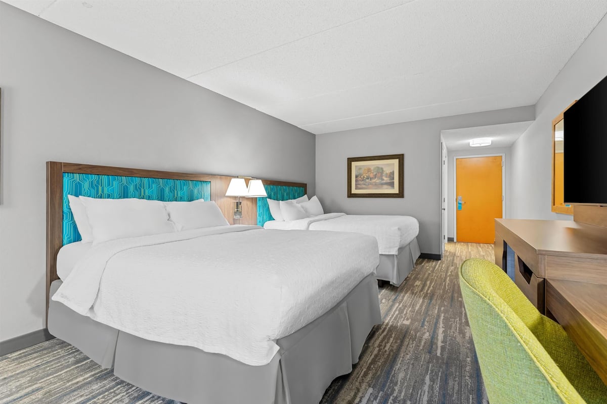 A hotel room with two beds, a desk, a chair, a lamp, and a painting on the wall.