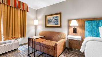Habitació d'hotel amb un sofà, una taula petita, una làmpada, un quadre a la paret, una finestra amb cortines, un radiador i un llit amb estriat blanc.