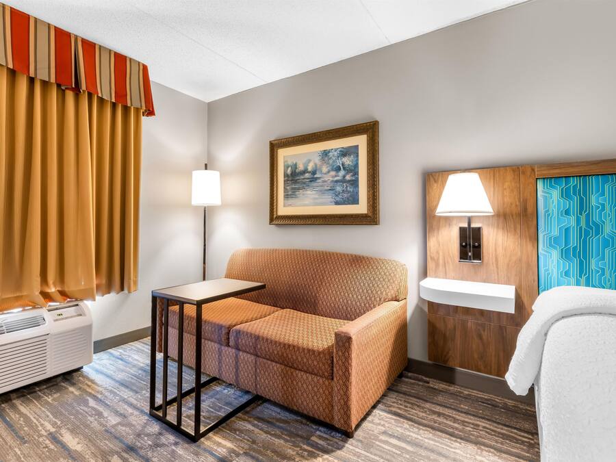 Habitació d'hotel amb un sofà, una taula petita, una làmpada, un quadre a la paret, una finestra amb cortines, un radiador i un llit amb estriat blanc.