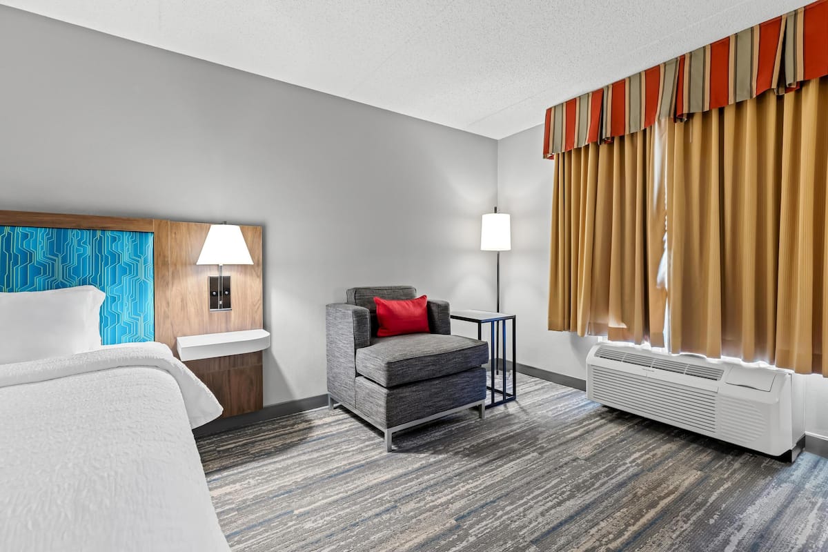 A hotel room with a bed, a gray armchair with a red pillow, a small table, a lamp, a window with curtains, and a wall-mounted heater.