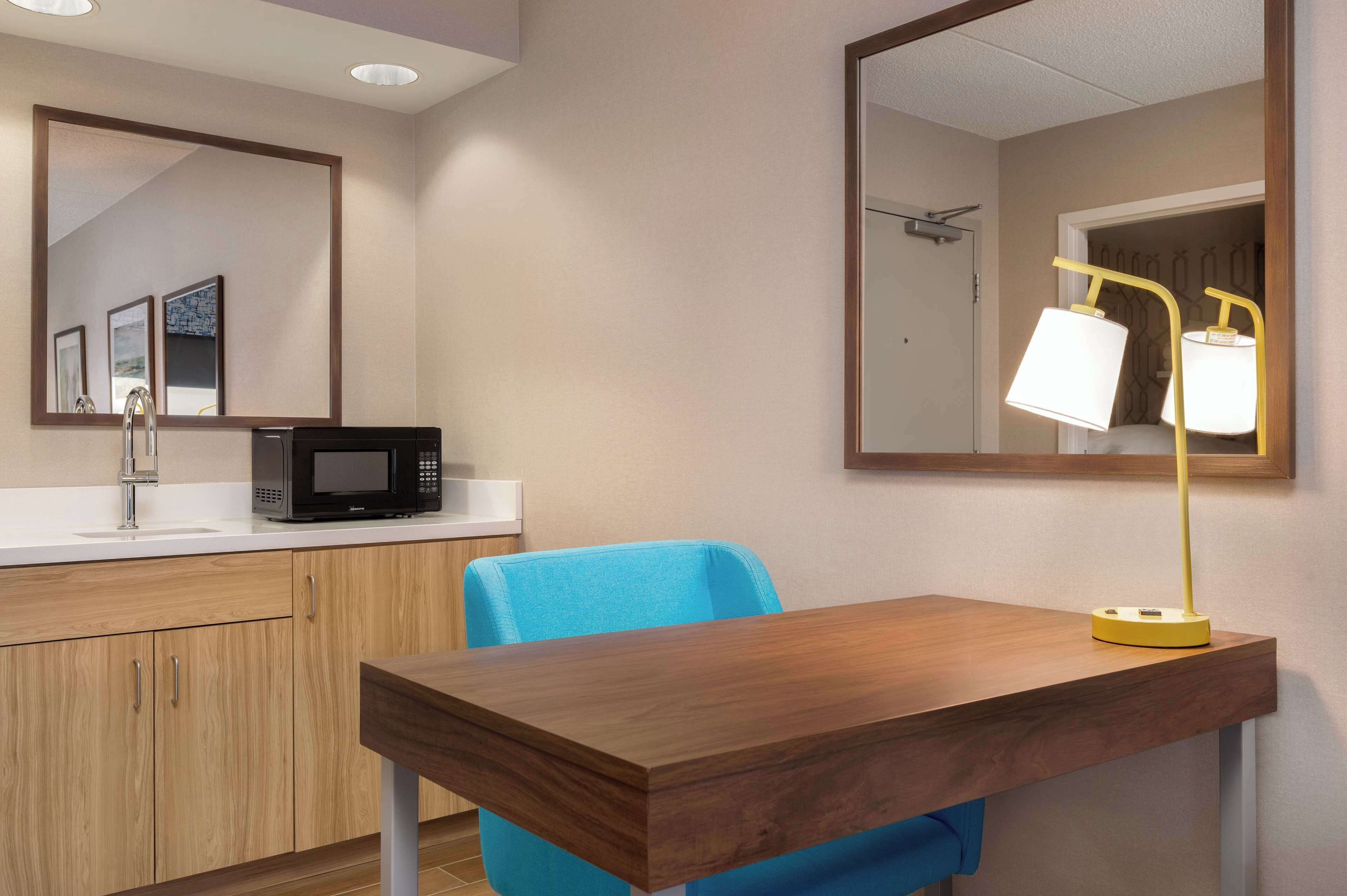 A compact kitchenette with a microwave, sink, and wooden cabinets. A blue chair and a wooden table with a yellow lamp are also visible.