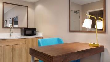 A compact kitchenette with a microwave, sink, and wooden cabinets. A blue chair and a wooden table with a yellow lamp are also visible.