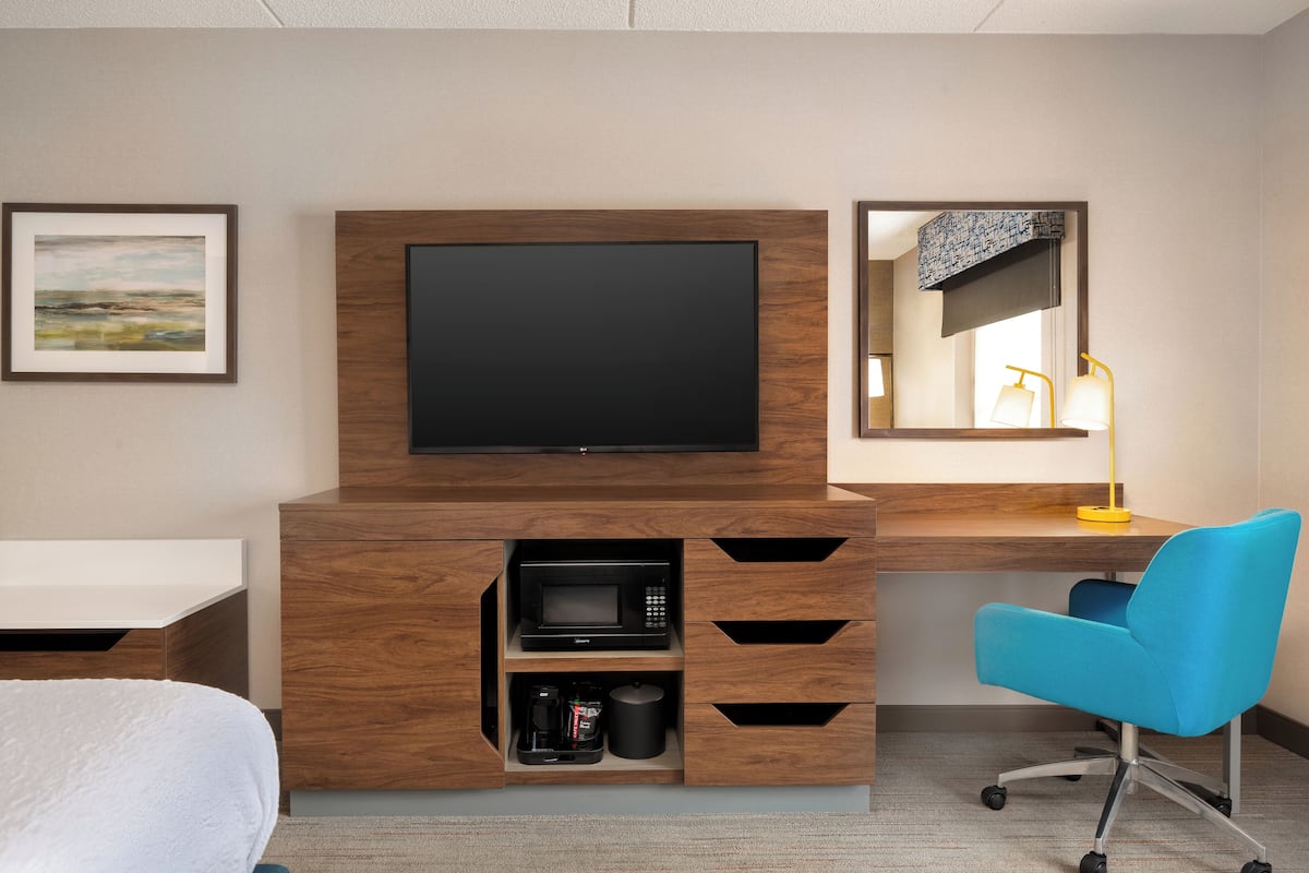 A hotel room with a wooden TV stand, a flat-screen TV, a microwave, a coffee maker, a desk, and a blue chair.