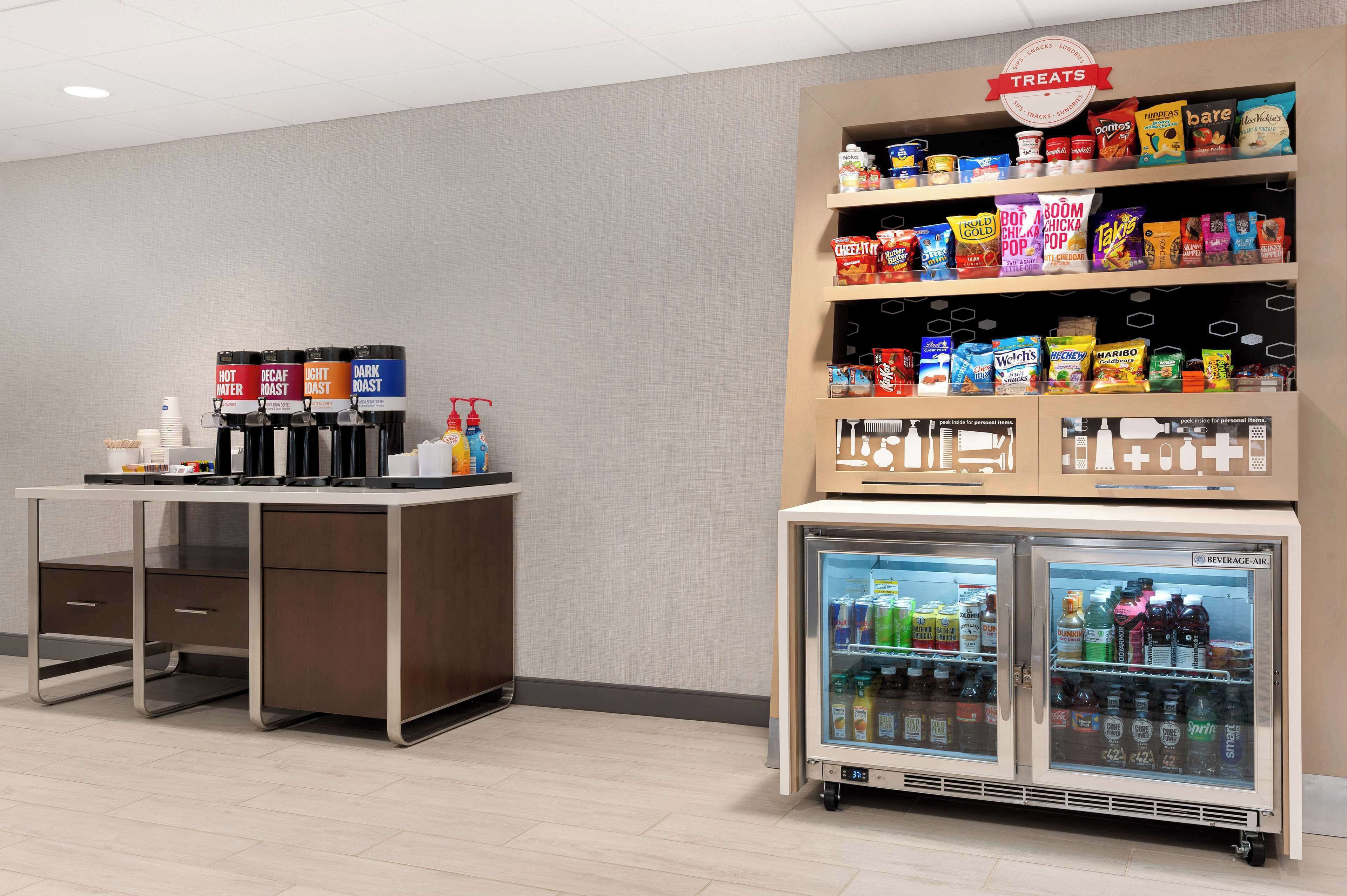 A self-service beverage station with hot, decaf, light roast, and dark roast options, a snack shelf with various brands, and a refrigerated drink cooler.
