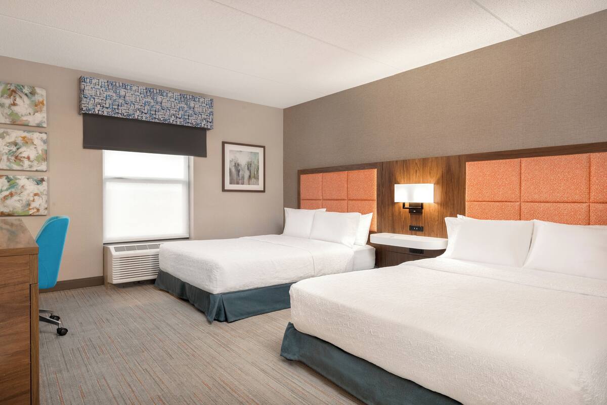 A hotel room with two beds, a desk, a chair, a window, and artwork on the walls.