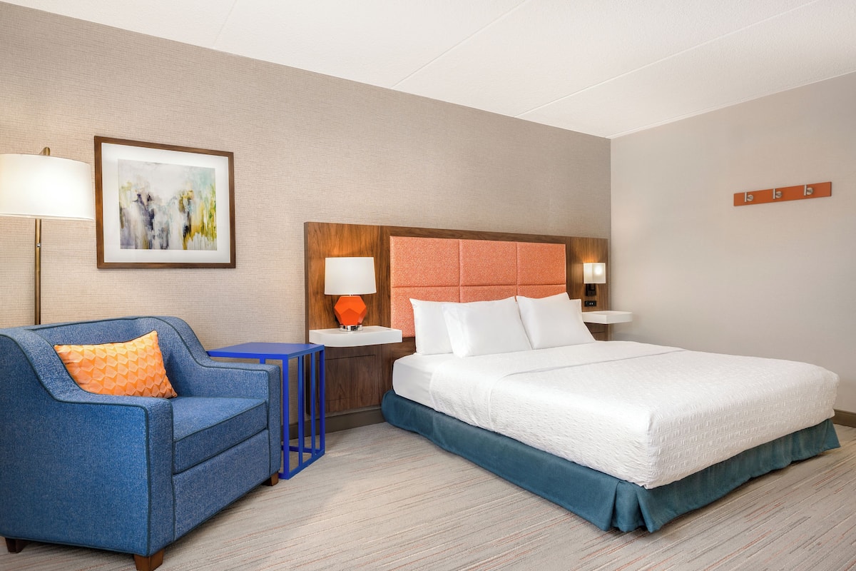 A hotel room with a bed, a sofa, a nightstand, a lamp, and a painting on the wall.