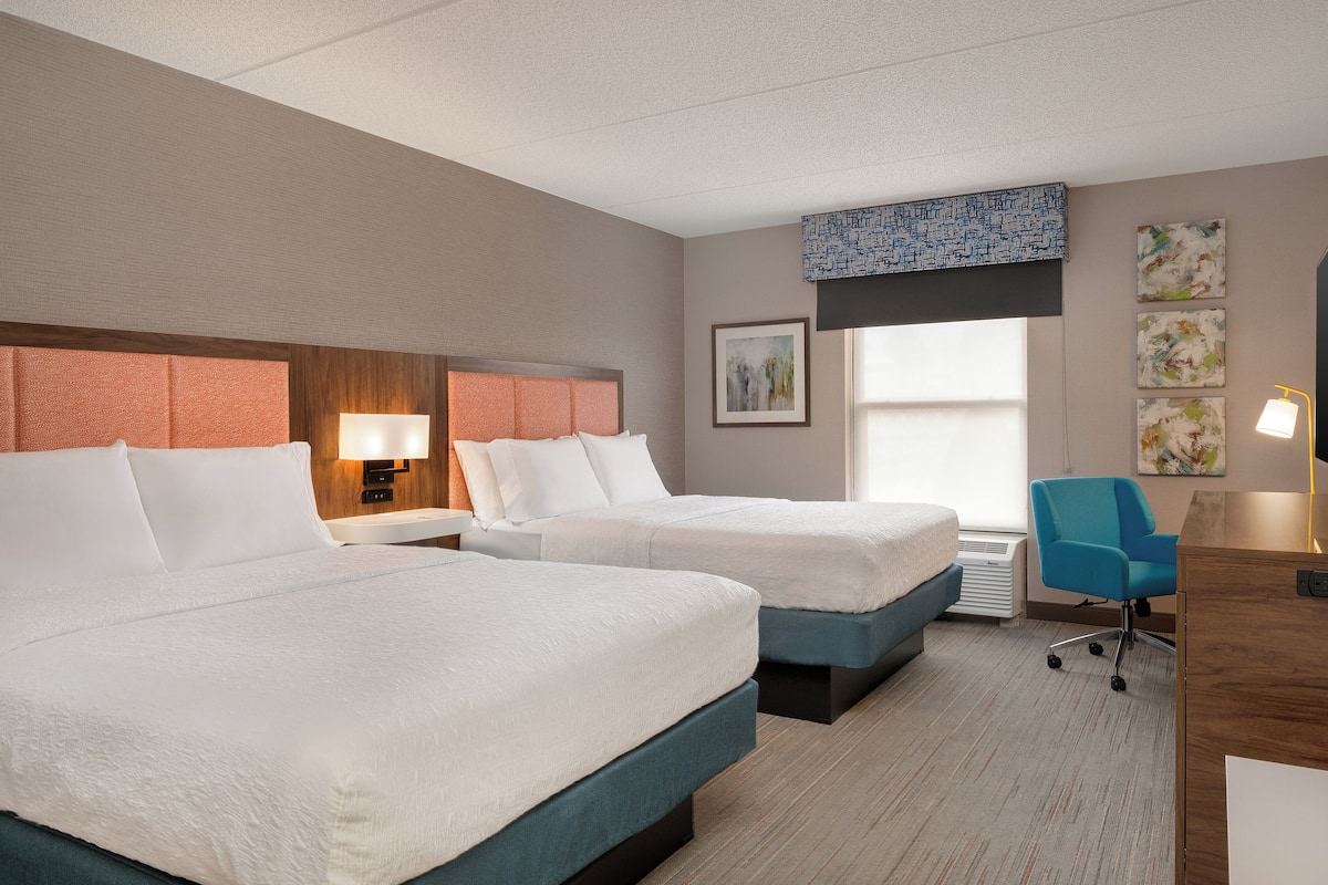 A hotel room with two beds, a desk, a chair, and artwork on the walls.