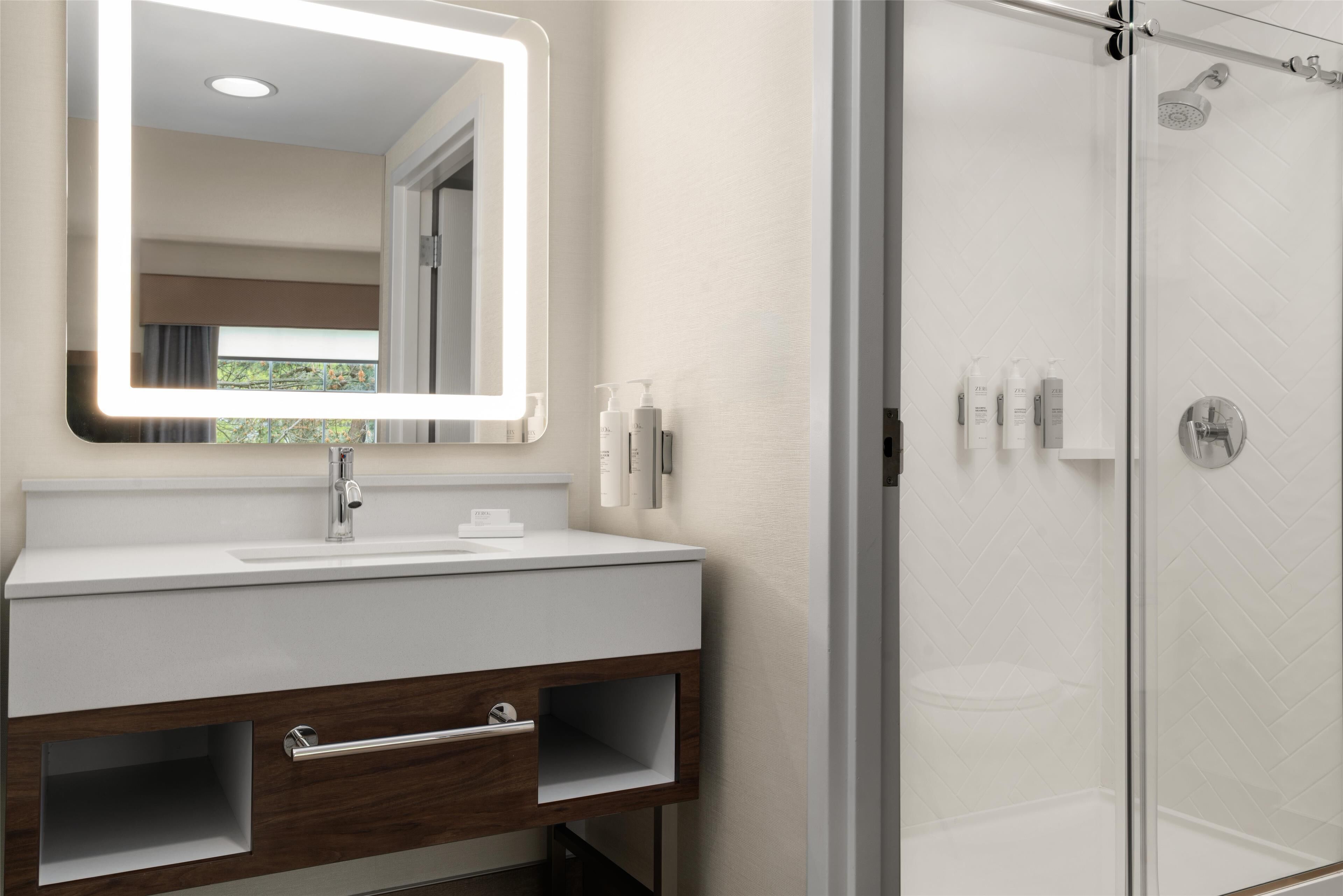 A modern bathroom with a white vanity, a large mirror with a light, and a glass-enclosed shower.