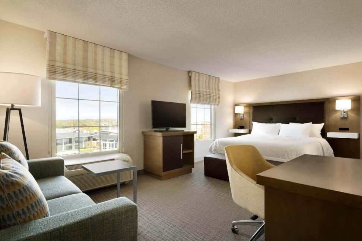 Habitación de hotel con cama, televisión, escritorio, silla, sofá y ventanas amplias.