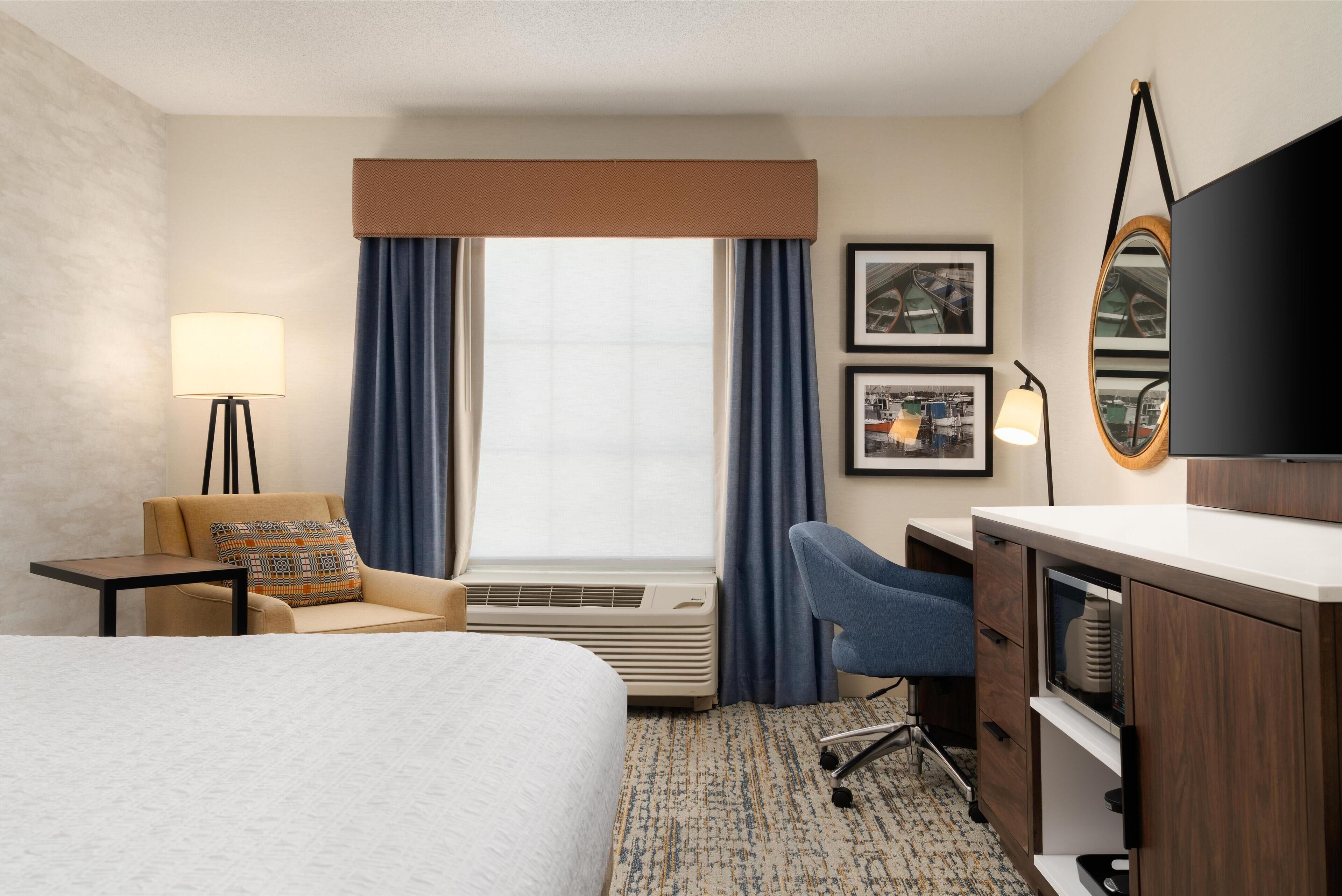 A hotel room with a bed, a desk, a chair, a lamp, a TV, and a window with curtains.