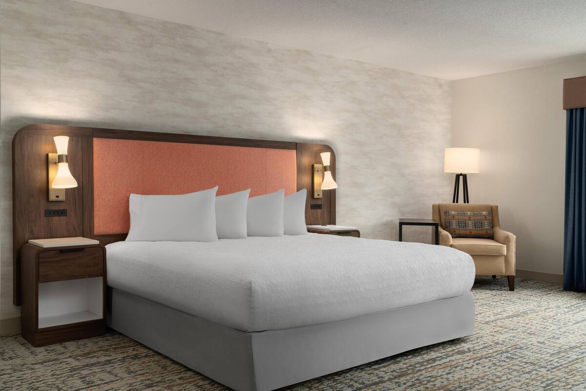 Habitación de hotel con una cama grande, dos mesitas de noche, una silla y una lámpara.