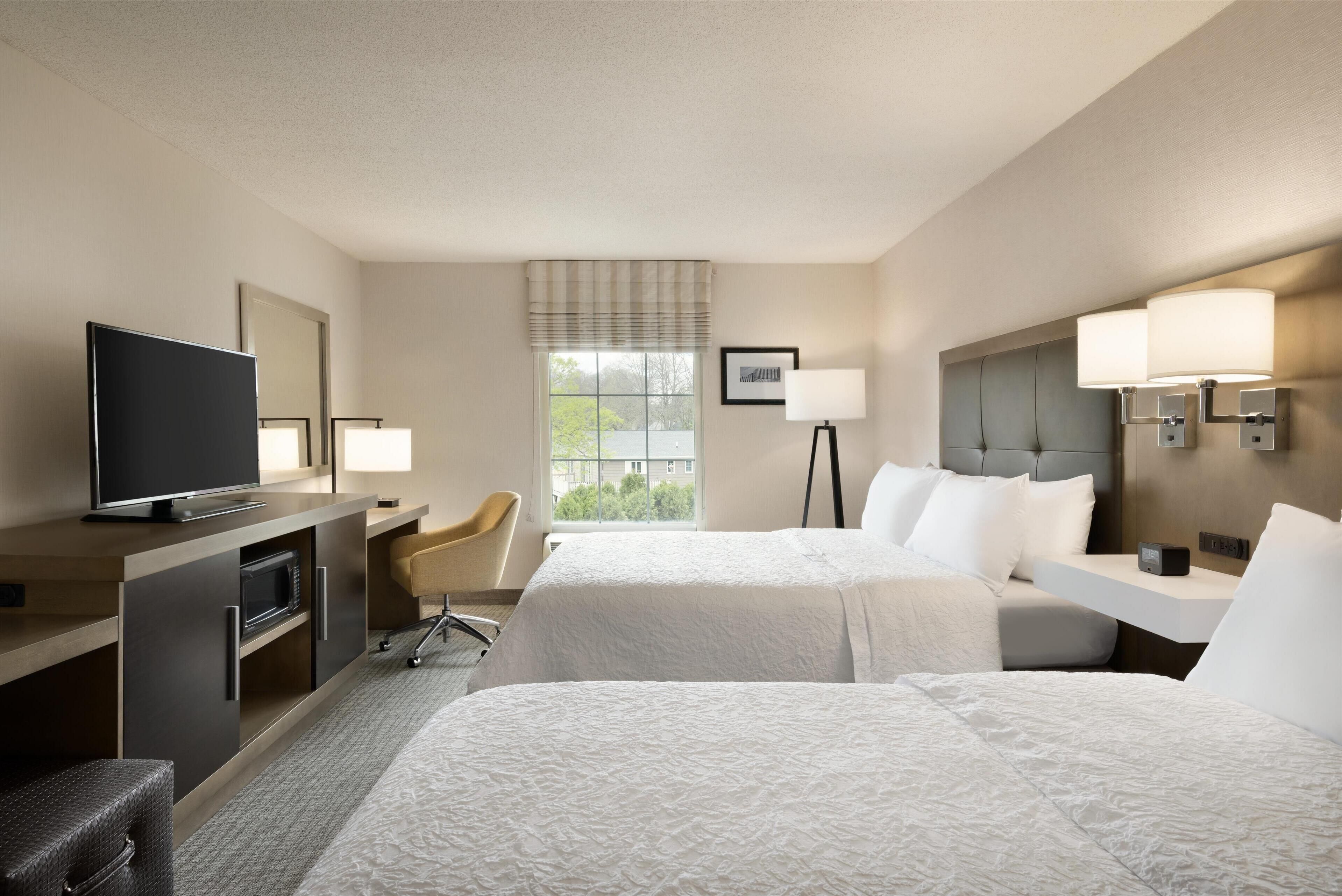 A hotel room with two beds, a desk with a television, a chair, and a window with a view of trees.