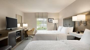 A hotel room with two beds, a desk with a television, a chair, and a window with a view of trees.